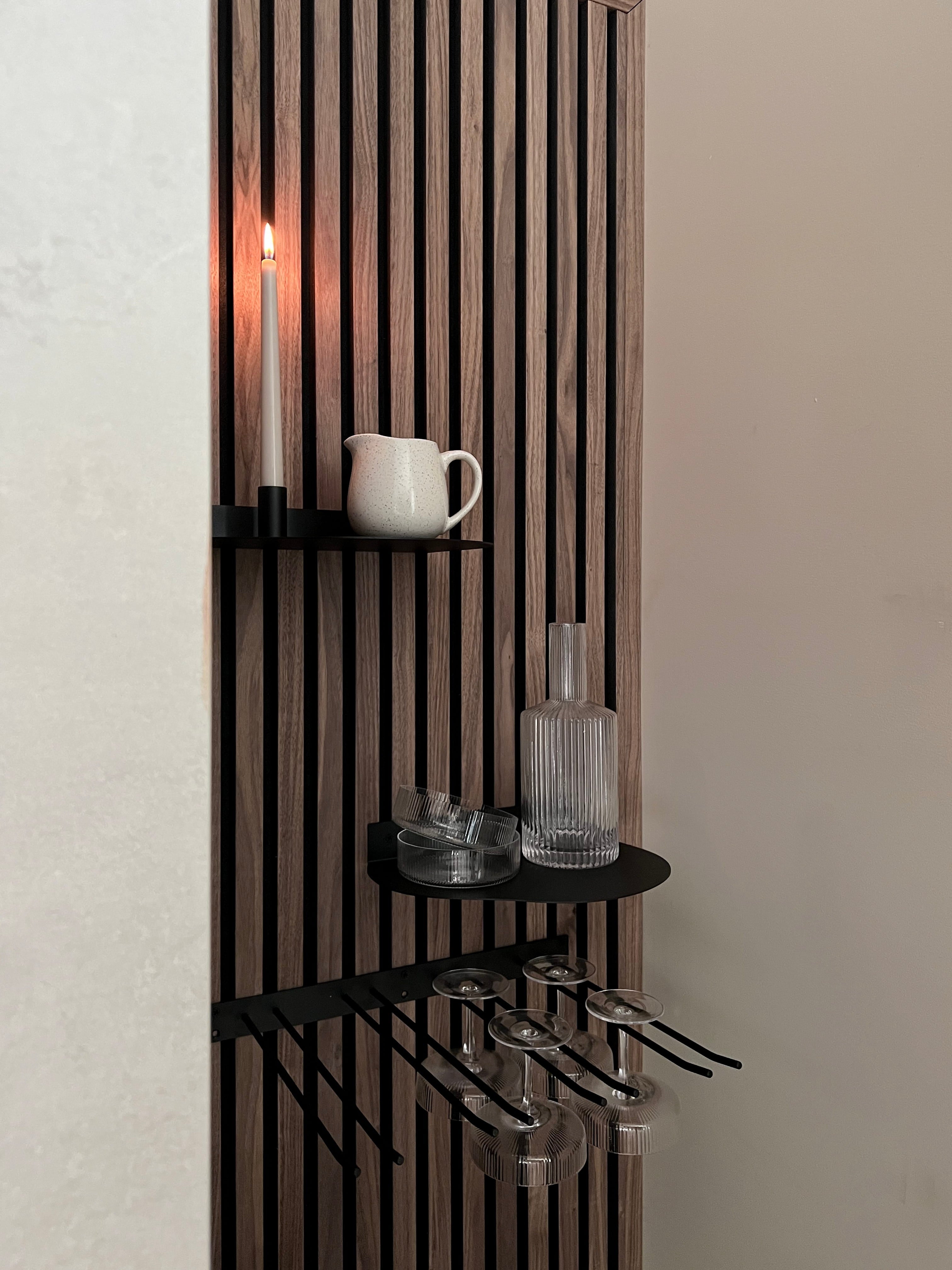 Painel acústico nórdico moderno de parede de ripas com prateleiras pretas contendo uma vela acesa, jarra branca, garrafa de vidro e taças de vinho com haste penduradas abaixo.